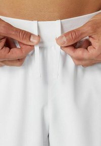 Manos ajustando los cordones blancos de la cintura de unos pantalones blancos contra un fondo de piel desnuda.