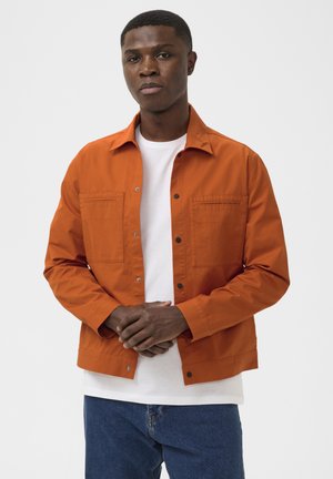Jeune homme portant une veste orange boutonnée par-dessus un t-shirt blanc et un jean bleu, debout les mains jointes devant lui.