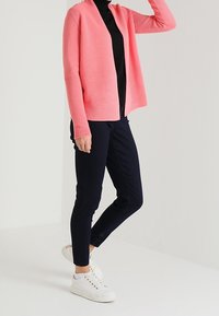 Koronkowy kardigan w kolorze koralu noszony rozpięty na czarnym golfie, w połączeniu z granatowymi spodniami taper i białymi trampkami.