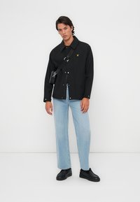 Veste noire à boutons-pression, col pointu et logo brodé ; associée à un jean bleu clair et des chaussures noires. Sac bandoulière réglable.