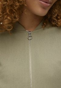 Oliwkowa kurtka zapinana na zamek z dopasowaną linią szyi, z metalowym zamkiem błyskawicznym z okrągłym uchwytem. Gładka struktura materiału.