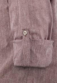 Chemise à manches longues en tissu mauve doux et texturé. Présente un poignet roulé avec un bouton en argent et un détail de couture près de l'ouverture du bouton.