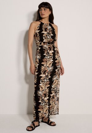 Frau trägt ein ärmelloses schwarzes Maxikleid mit beigen und braunen Blattmustern, tailliertem Gürtel und schwarzen Riemchensandalen, steht an einer Wand.
