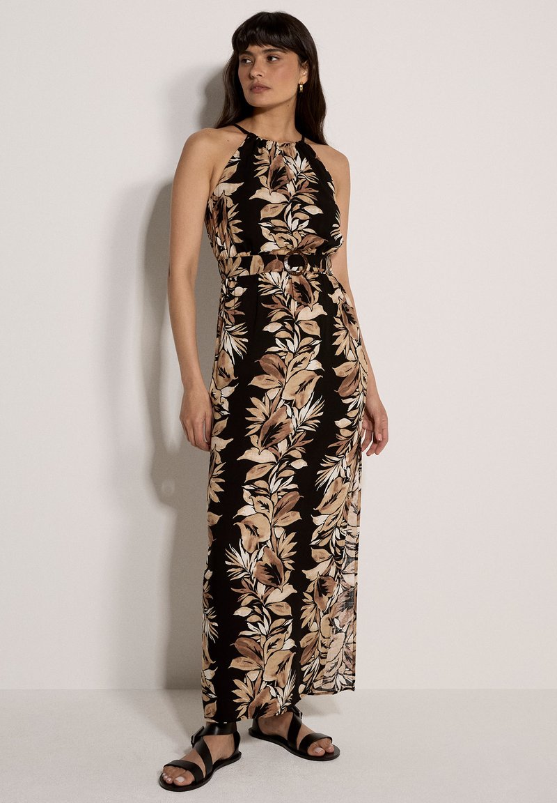 Femme portant une robe maxi noire sans manches avec des motifs de feuilles beiges et marron, ceinte à la taille, et des sandales noires à lanières, debout contre un mur.