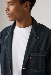 Σκούρο μπλε τζιν σακάκι με γιακά σε σχήμα V, μία μπροστινή τσέπη και κίτρινη ραφή. Φορεμένο πάνω από ένα λευκό μπλουζάκι με στρογγυλή λαιμόκοψη.