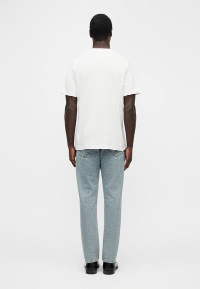 Valge puuvillane t-särk, lühikeste varrukatega, lõdva istuvusega, kombineeritud hele siniste teksapükstega. Lihtne disain, tavaline tagantvaade, nähtavad mustrid või aktsendid puuduvad.