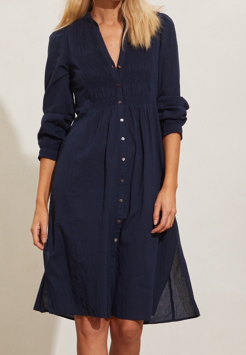 Femme portant une robe boutonnée bleu marine avec des manches longues retroussées et des détails plissés subtils sur la poitrine.
