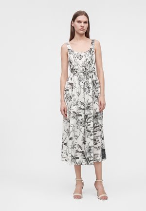 Femme portant une robe midi blanche sans manches avec un imprimé floral noir et des talons clairs à bride à la cheville, debout devant un fond uni.