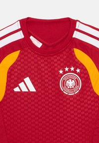 Κόκκινη αθλητική φανέλα με λευκές και κίτρινες λεπτομέρειες, με υφασμάτινο σχέδιο, στρογγυλή λαιμόκοψη και το λογότυπο της Γερμανικής Ποδοσφαιρικής Ομοσπονδίας με αστέρια.