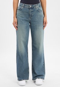 Jasnoniebieskie jeansy szerokie w nogawkach, wykonane z denimu, o czystym, wyblakłym wzornictwie z zapięciem na guzik i pięcioma kieszeniami. Widać białe trampki.