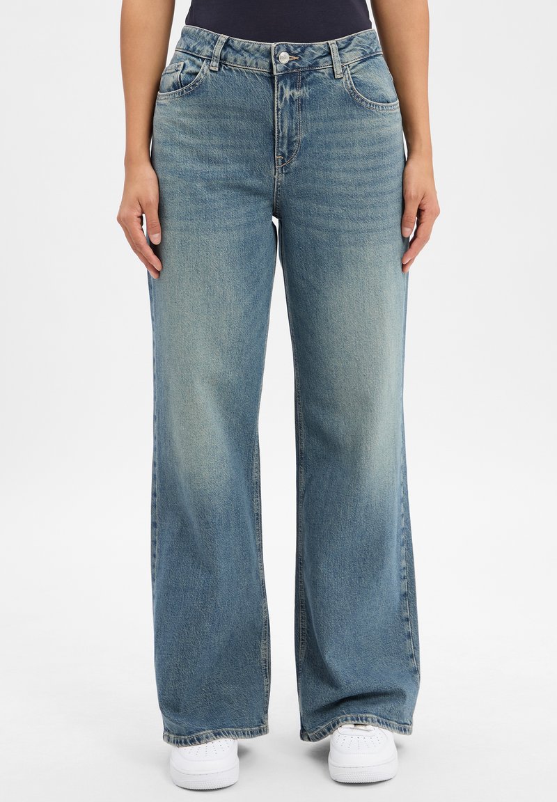 Jasnoniebieskie jeansy szerokie w nogawkach, wykonane z denimu, o czystym, wyblakłym wzornictwie z zapięciem na guzik i pięcioma kieszeniami. Widać białe trampki.