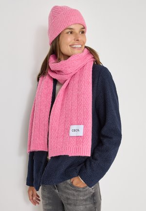 Sciarpa rosa lavorata a maglia con un motivo a spina di pesce, esposta con un cappello abbinato. La sciarpa presenta un'etichetta di marca ed è realizzata in materiale morbido.