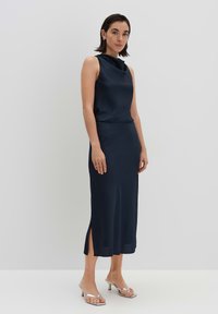 Dunkelblaues ärmelloses Kleid mit einem Cowl-Ausschnitt, strukturiertem Stoff und seitlichem Schlitz; kombiniert mit silbernen Riemchensandalen. Schlichtes, figurbetontes Design.