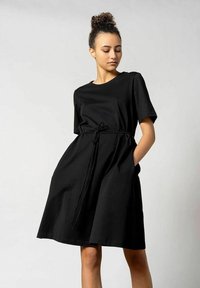 Schwarzes Kurzarmkleid aus weichem Stoff mit lockerer Passform, einem Zugband in der Taille und seitlichen Taschen. Glatte Textur und knielanges Design.