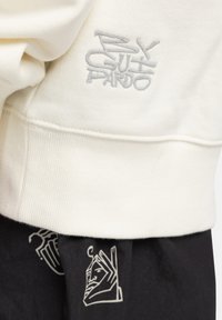 Cream sweatshirt with gray embroidered logo on the hem; paired with black fabric featuring a white abstract face pattern.