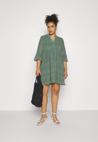 Grünes, kurzes Kleid mit Spitzenverzierungen und V-ausschnitt. Kombiniert mit einer schwarzen, geflochtenen Tasche und weißen, schmalen Sandalen.