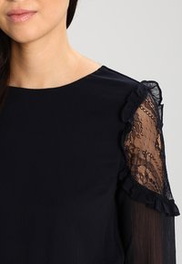 Námorně modrá blůza s krajkovými detaily na rukávech, má volánové okraje a hladkou texturu látky, nabízí volný střih a zaoblený výstřih.