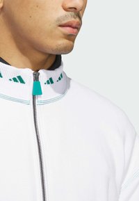 Giacca bianca con zip, caratterizzata da un alto collo con dettagli in teal e texture a coste. La zip silver contrasta con il tessuto.