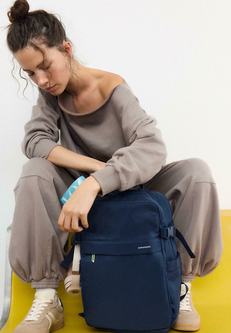 Femme en tenue de détente taupe assise, tenant un sac à dos bleu avec un magazine plié à l'intérieur, sur un fond blanc et jaune.