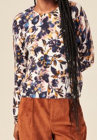 Valkoinen kukallinen neulepaita, jossa sinisiä, keltaisia ja vaaleanpunaisia kukkia; pyöreä pääntie ja ribboitettu helma; yhdistetty ruskeisiin corduroy-housuihin.