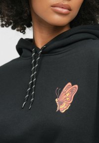 Czarna bluza z kapturem z grafiką motyla w odcieniach pomarańczowego i żółtego. Teksturowany materiał z czarnymi i białymi sznurkami do ściągania.