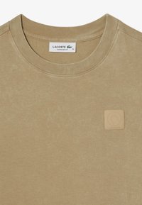 Camiseta oversize beige de tela suave con cuello redondo. Presenta un parche de cuero texturizado con el nombre de Lacoste en la esquina inferior.