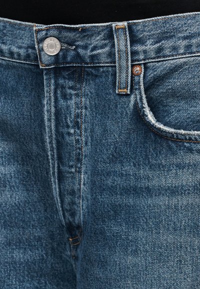 Blaue Jeans aus Denim mit Knopfverschluss, tonalem Steppmuster und einem leicht ausgefransten Rand an der Tasche, der einen lässigen, getragenen Look betont.