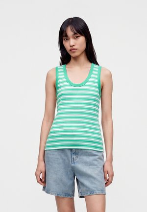 Kvinde iført en grøn- og hvidstribet ærmeløs top og lyseblå denimshorts, stående mod en ensfarvet lys baggrund.