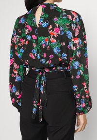 Blusa preta com padrões florais vibrantes em rosa, azul e verde; possui cintura franzida, mangas longas e detalhe de abertura em forma de gota nas costas.
