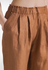 Nærbillede af talje og hånd i lomme iført løstsiddende, højtaljede, brune hørbukser med elastisk, rynket talje og forlommer med læg.