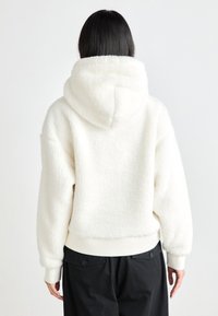 Faux fur white hoodie with a hood, ribbed cuffs, and a short hem. Features a soft, textured exterior and is paired with black pants.