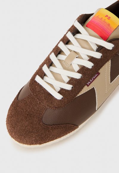 Sneaker din suede și piele maronie cu șireturi albe, având un accent bej și un eticheta multicoloră la limbă. Design cu vârf rotund.