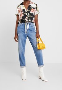 Blommig svartvit skjorta med knappar knuten i midjan, ihop med ljusblå högmidjade jeans och vita ankelboots. Gul handväska.