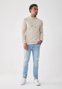 Hoodie bege em malha com cordão, combinada com jeans azul claro. A modelo usa ténis brancos e apresenta uma silhueta ajustada.