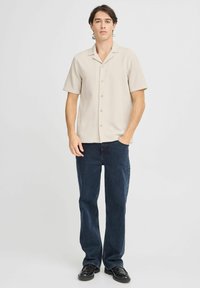 Camisa de botones de manga corta de color beige claro, con tejido texturizado. Combinada con jeans azul oscuro y zapatos negros. De pie en una pose relajada.
