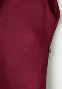 Primo piano di un indumento in tessuto color granata che mostra una cerniera laterale parzialmente visibile con cursore abbinato su uno sfondo uniforme.