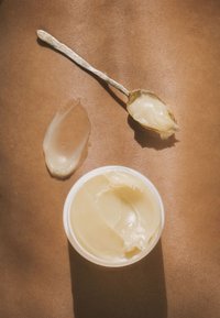 Krämig beige kroppskräm i en vit behållare, med en silver sked och en klick på en jämn, solbränd yta. Mjuk textur, glansig finish.