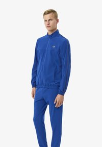 Uomo in piedi che indossa una giacca a zip blu e pantaloni coordinati, con un piccolo logo di coccodrillo sulla parte superiore del petto.
