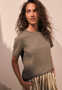 T-shirt verde oliva a maniche corte in tessuto morbido, abbinata a gonna a righe e plissettata di colore chiaro. Texture liscia e vestibilità rilassata.