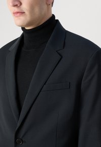 Blazer nero con rever appuntito, bottone singolo e taschino sul petto. Indossato su un dolcevita nero, che mette in evidenza una texture liscia.