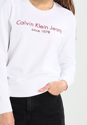 Sweatshirt blanc à manches longues avec logo brodé rouge "Calvin Klein Jeans depuis 1978" ; poignets et ourlet côtelés. Texture de tissu uni.
