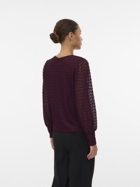 Kvinne med hår i bun som har på seg en mørk lilla teksturert langermet bluse og svarte bukser, står med ryggen mot en enkel hvit bakgrunn.