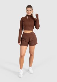 Brun kort zip-up jacka som matchar med shorts. Båda är tillverkade av elastiskt tyg. Har hela ärmar och en avslappnad passform.