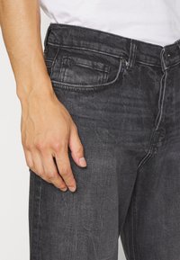 Jeans in denim nero a vestibilità slim, con cuciture visibili, tasche anteriori e posteriori, e una trama sottile. Mano posizionata sulla coscia.