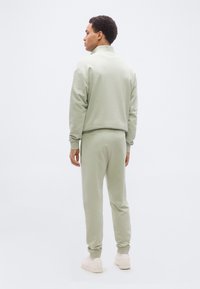 Άνδρας που στέκεται με την πλάτη προς τα εμάς, φοράει ανοιχτοπράσινο φούτερ και τζόγγερ που ταιριάζουν με λευκά αθλητικά παπούτσια, σε απλό λευκό φόντο.