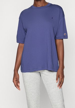 Person iført en løs, kortærmet, blå Champion t-shirt og lysegrå tætsiddende bukser mod en ensfarvet baggrund.