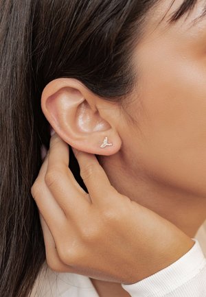 Rose gold ear cuff with small, clear gemstones, shaped like a stylized wing, worn on a model's ear. Skin tone and hand visible.