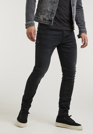 Jeans ajustados de mezclilla negra con un acabado desgastado, combinados con una chaqueta de mezclilla gris y zapatillas negras con una parte superior texturizada y suela blanca.