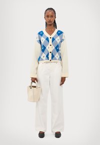 Cardigan bleu et blanc à motifs argyle avec des boutons, patte de boutonnage couleur crème, pantalon large blanc, et un petit sac à main beige. Chaussures noires.