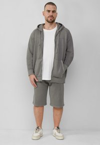 Lichtgrijze rits hoodie met voorzakken, gecombineerd met bijpassende shorts. Lichtgewicht stof; sneakers in beige met zwarte accenten.